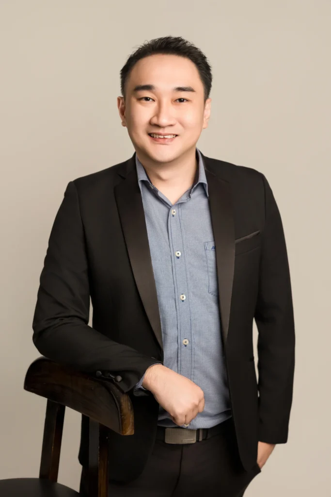 Professional portrait of Ar. Kenneth Fung, Founder and Principal Architect of M Interior Studio, leaning on a dark wood chair in a black blazer against a neutral studio backdrop.