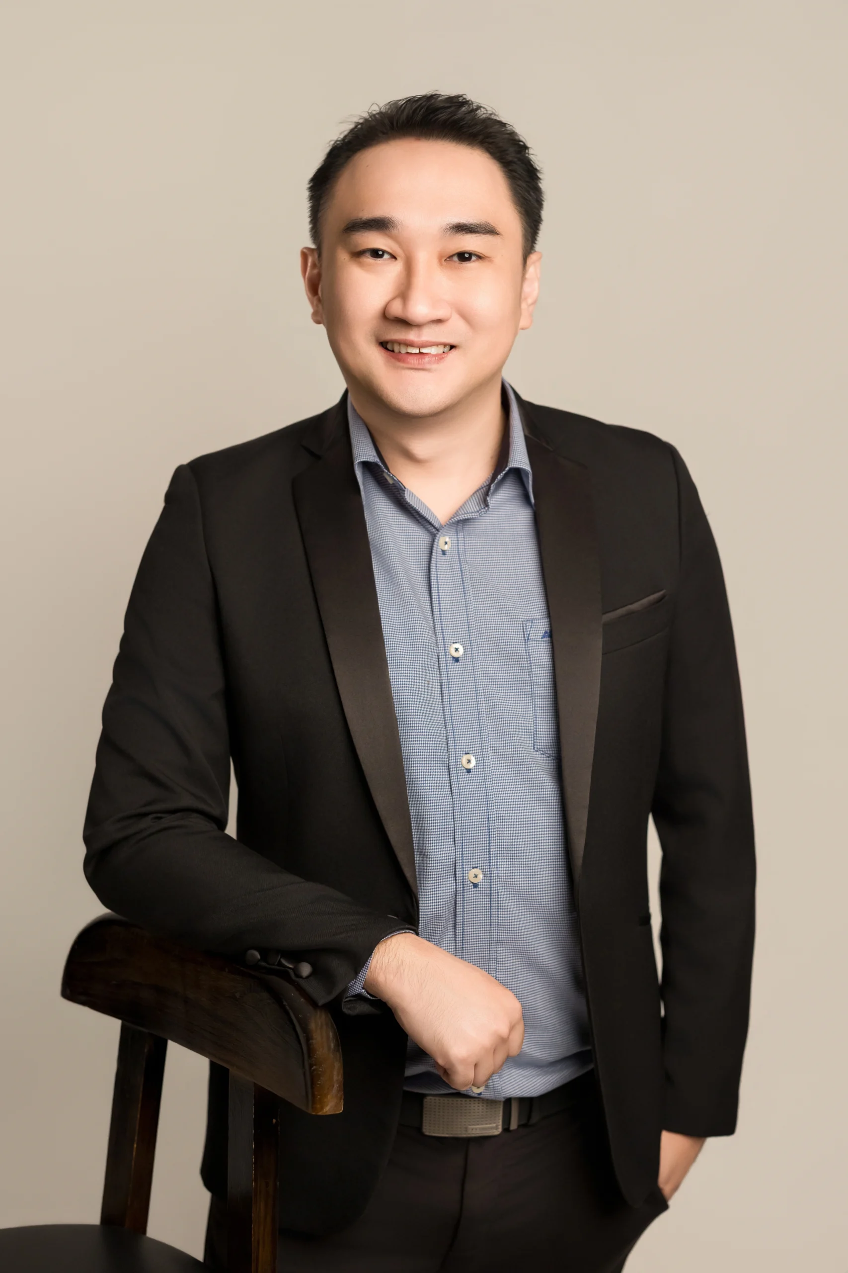Professional portrait of Ar. Kenneth Fung, Founder and Principal Architect of M Interior Studio, leaning on a dark wood chair in a black blazer against a neutral studio backdrop.