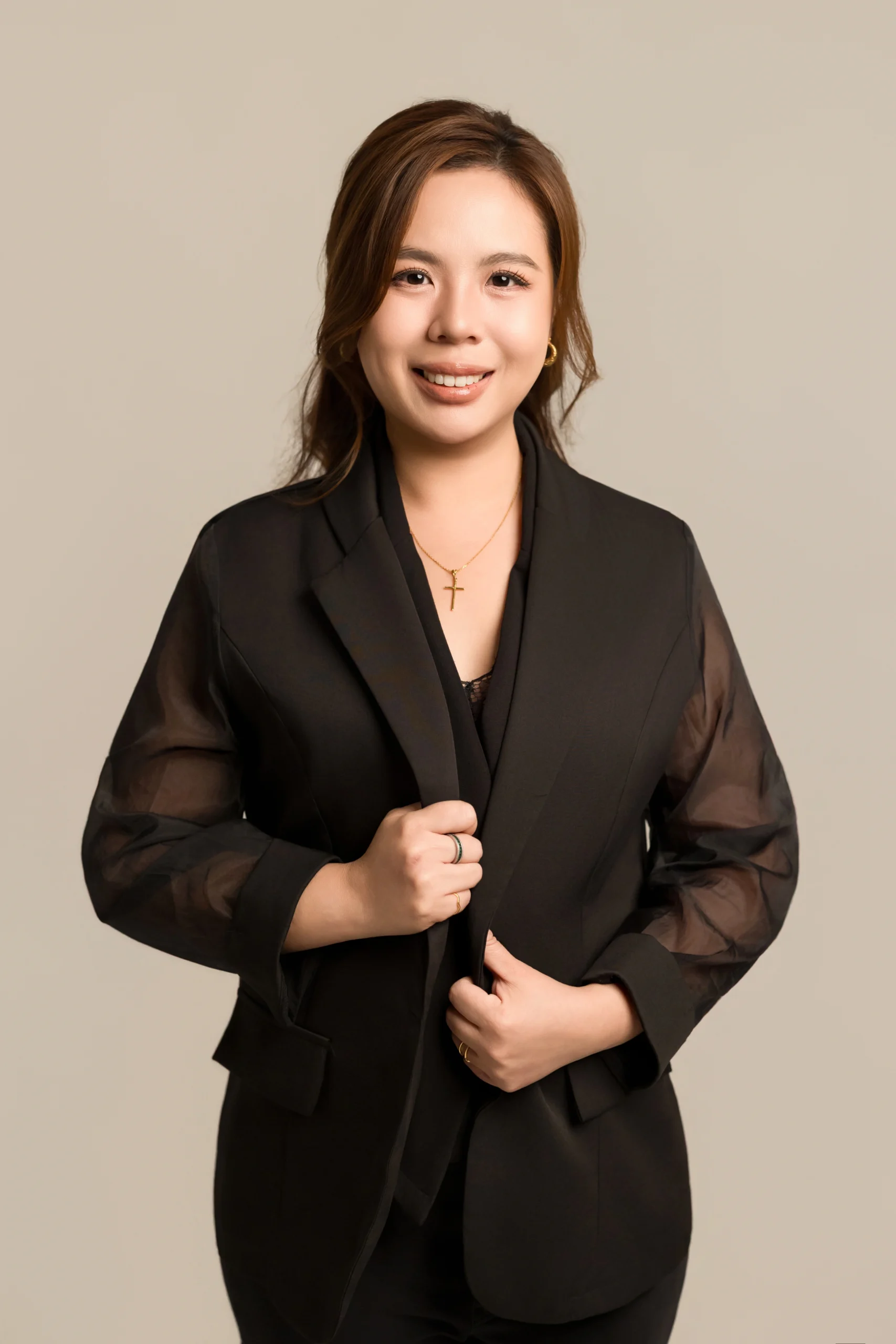 Professional headshot of Valencia Vu, CEO of M Interior Studio, wearing a stylish black blazer with sheer sleeves and a gold cross necklace, smiling against a warm neutral studio background.